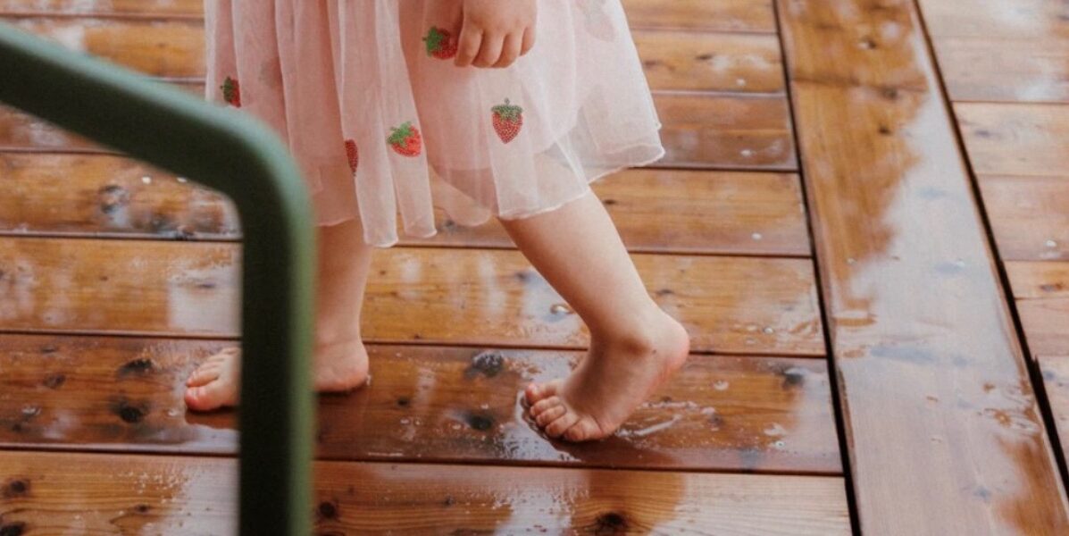 A child is walking on tiptoe on a wooden deck in the rain. She is wearing a skirt with a strawberry pattern.